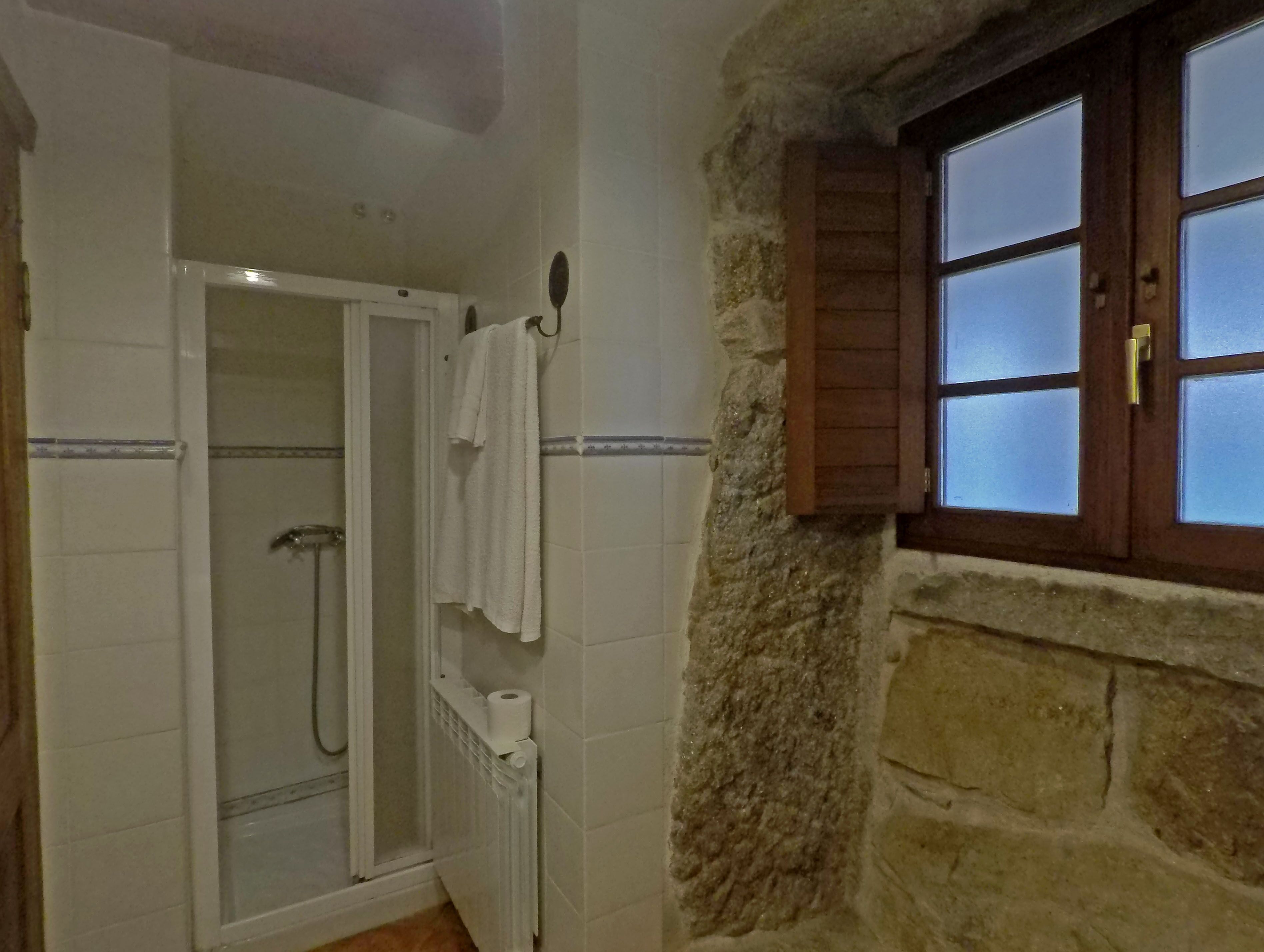 A bathroom with a glass-enclosed shower, a towel hanging on the wall, and a wooden window with a brass handle.