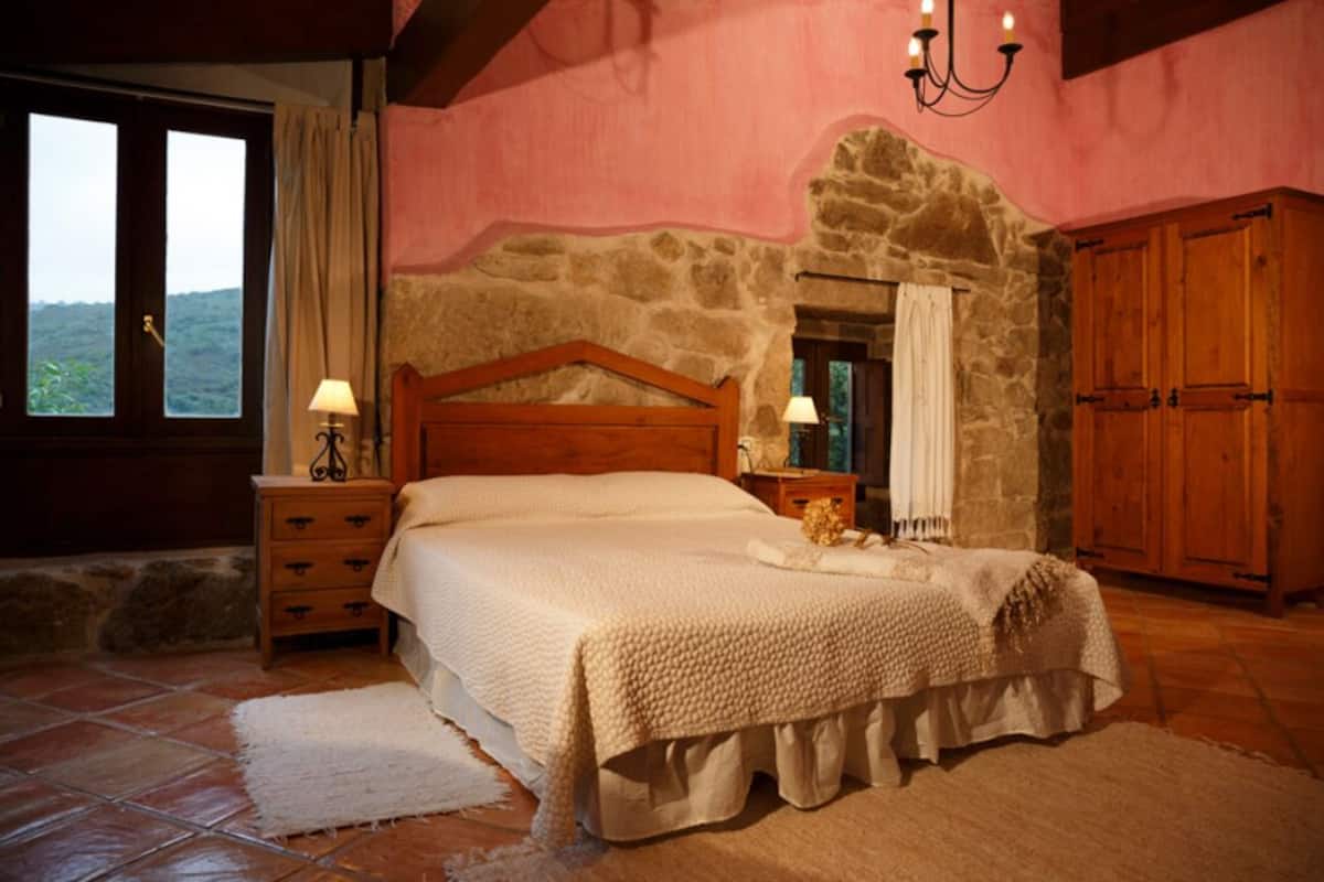 Quarto com parede de pedra, cama de madeira, mesa de cabeceira, guarda-roupa e janela com cortinas.