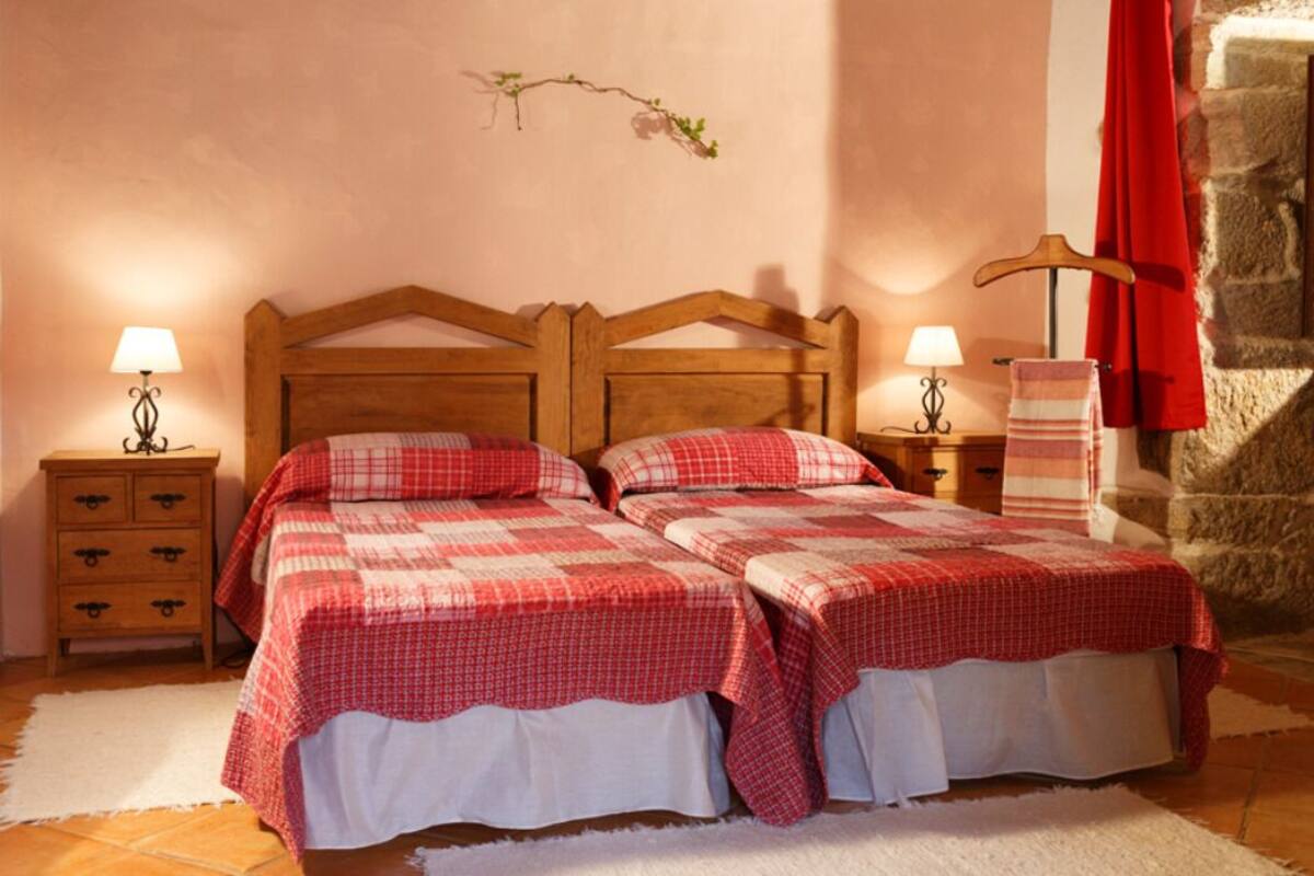 Uma cama de casal com cabeceira de madeira, duas mesas de cabeceira com abajures, um edredom xadrez vermelho e branco, um tapete branco e uma parede de pedra com decoração de galho.