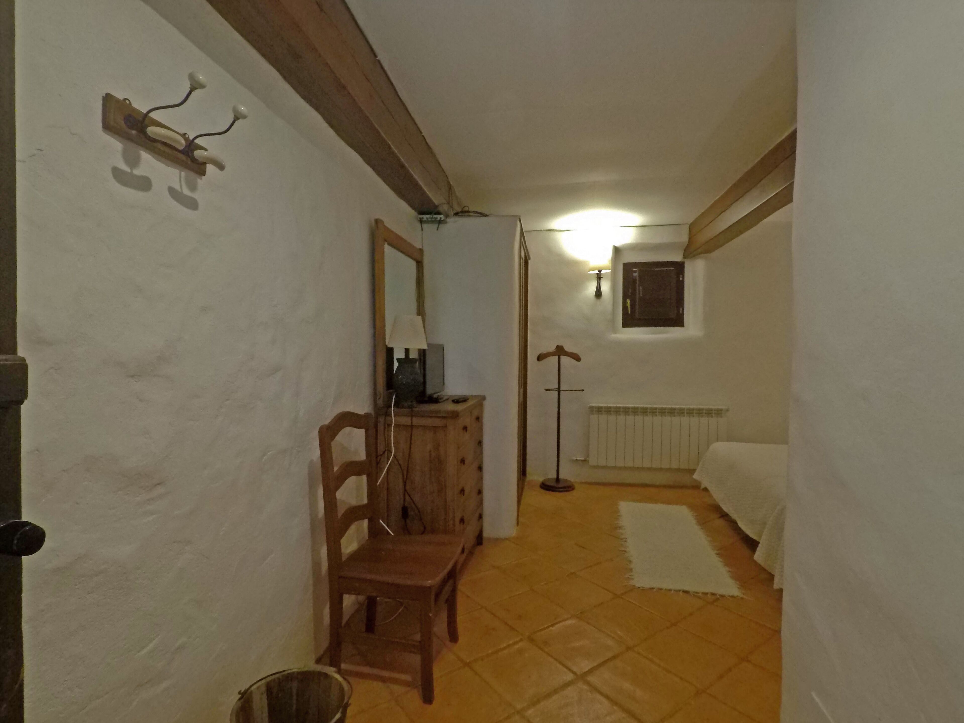 Un pasillo estrecho con una silla de madera, una mesita con una lámpara, un radiador y una ventana con persiana cerrada.