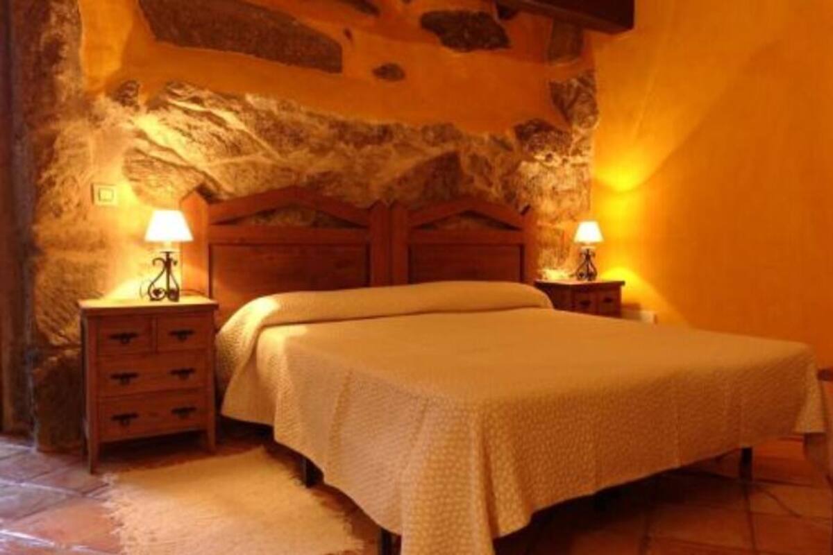 Quarto com parede de pedra, estrutura de cama de madeira, duas mesas de cabeceira com luminárias e uma cadeira.