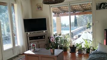 Een ruime woonkamer met een televisie, een groot raam en uitzicht op een zwembad en tuin.