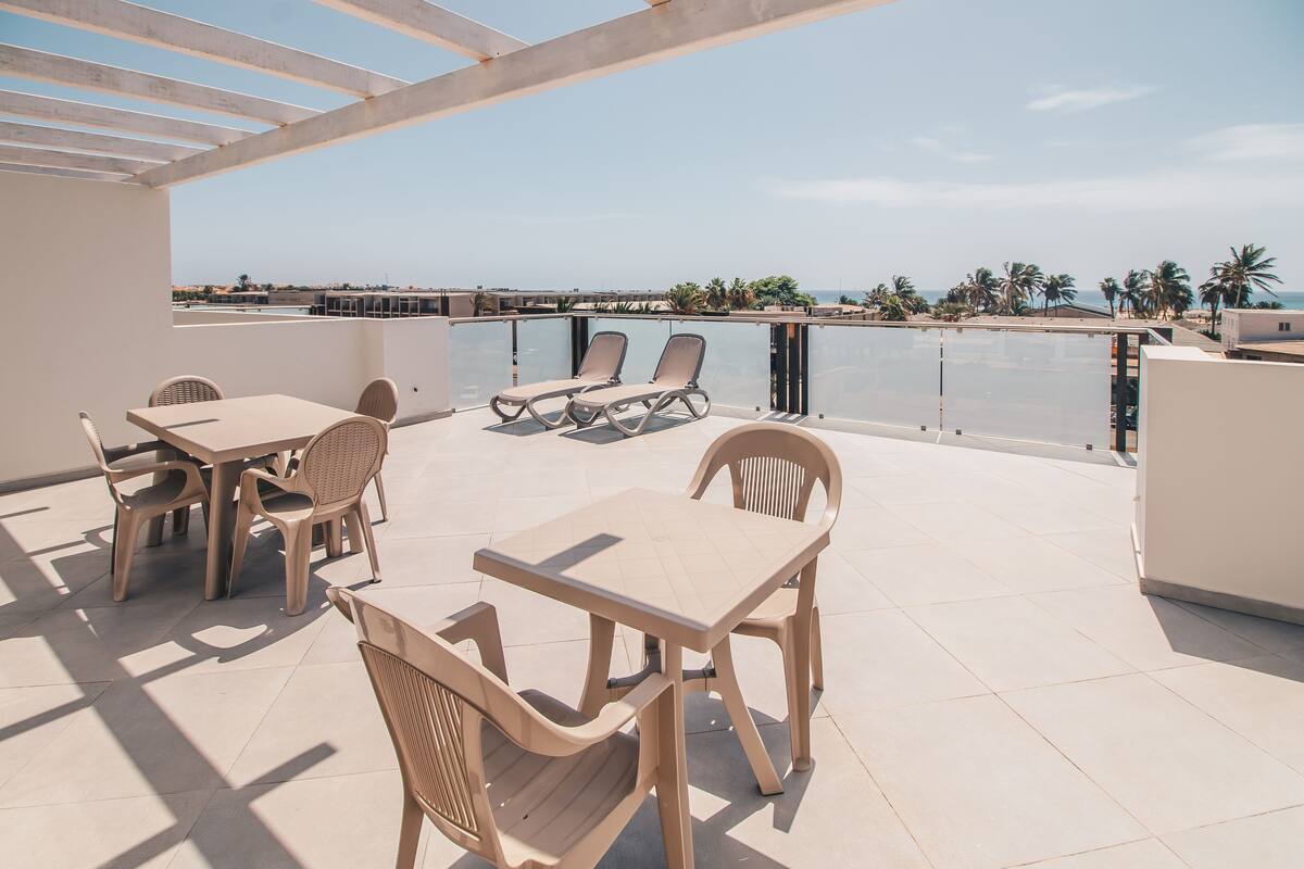 A terrace with a table and chairs, a sun lounger, and a view of the sea and palm trees.