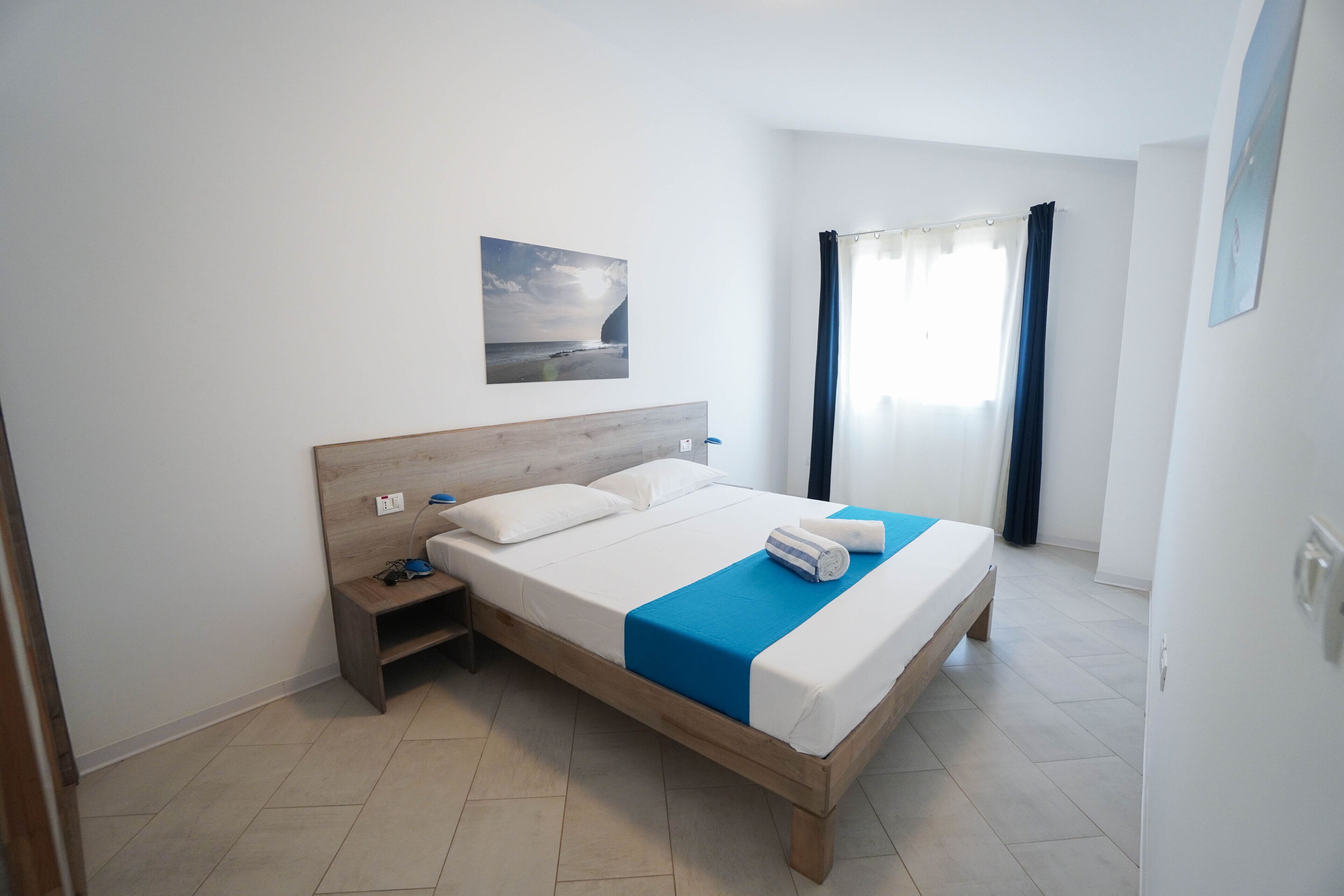 Une chambre d’hôtel moderne avec un lit en bois, une table de chevet, un tableau accroché au mur et une grande fenêtre avec des rideaux.