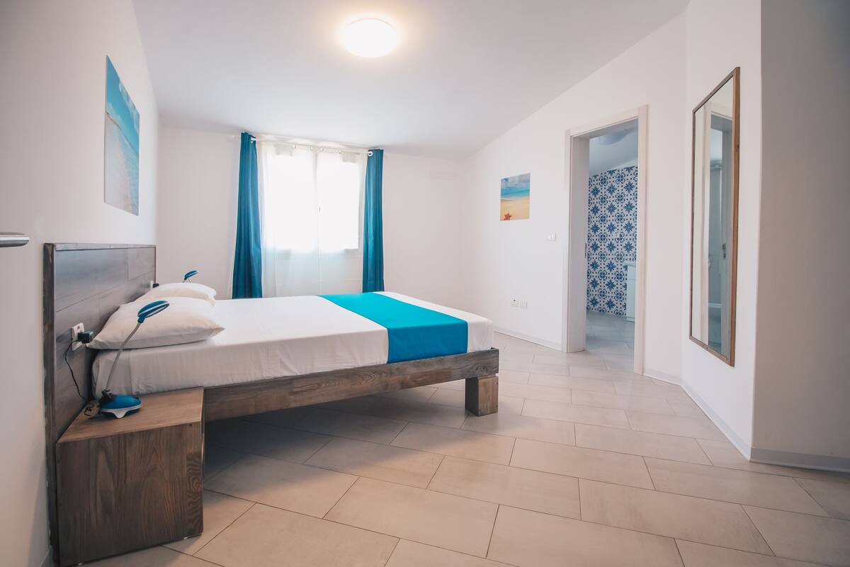 A bedroom with a wooden bed, a bedside table, a nightstand, a ceiling light, and a bathroom visible through an open door.