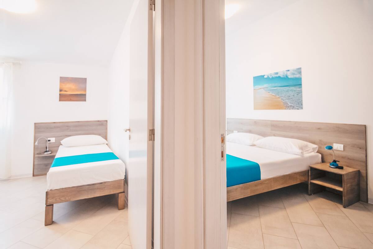 A hotel room with two beds, a nightstand with a lamp, a painting on the wall, and a view of the beach.