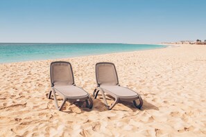Two empty lounge chairs on a sandy beach with a clear blue sky and calm sea in the background. - Halos Casa Resort (Sal)