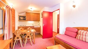 A compact living space with a kitchenette, a dining area with wooden chairs, a red door, and a sofa with patterned cushions. - Bel Appart proche du Centre, accès Sauna + Piscine (Valfréjus)