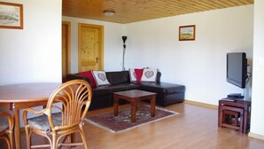 Una sala de estar con techo de madera, un sofá negro con cojines rojos, una mesa de centro de madera, un mueble para televisión, y una zona de comedor con mesa de madera y sillas de mimbre.