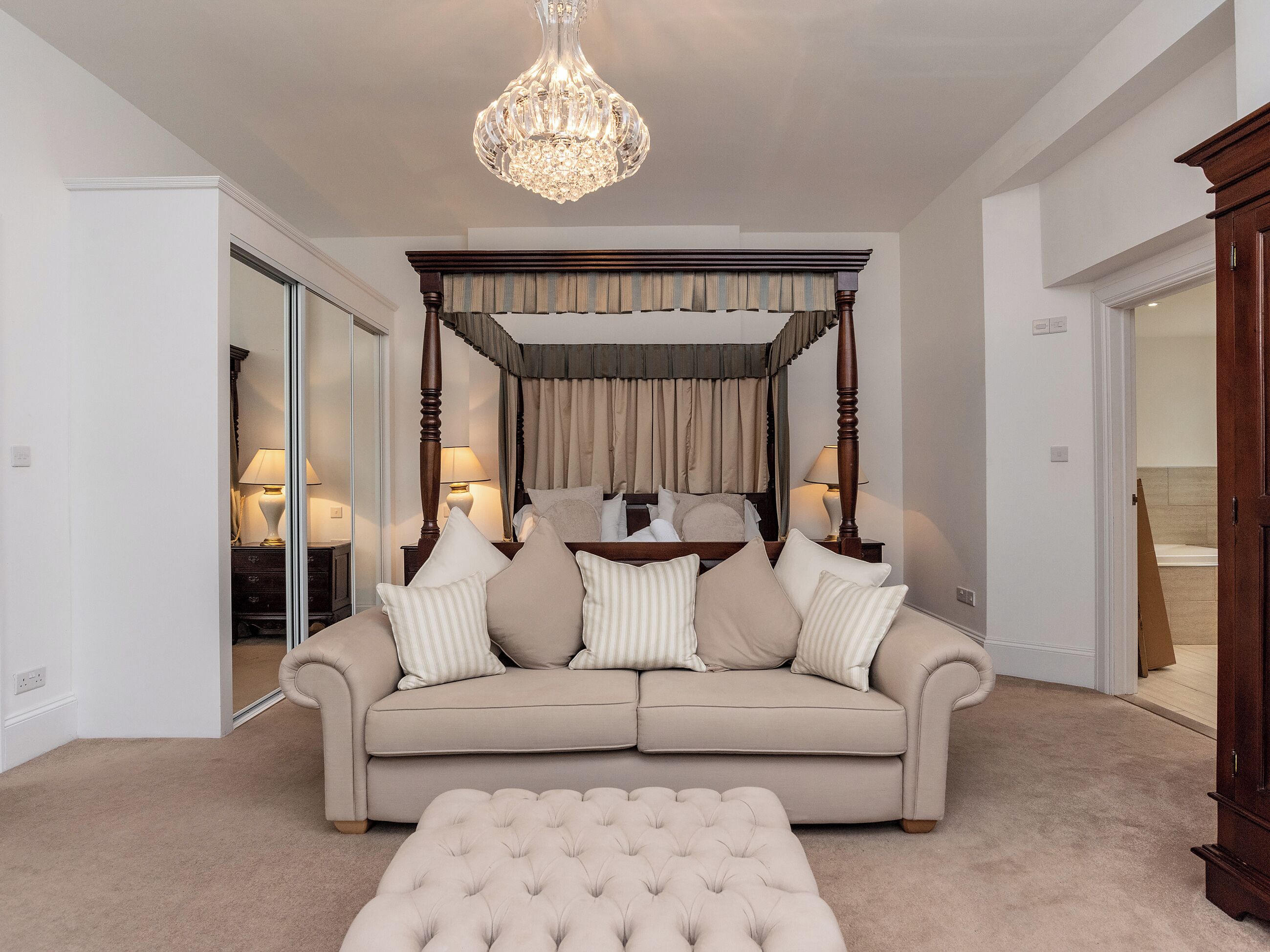 A bedroom with a four-poster bed, a beige sofa, and a tufted ottoman.