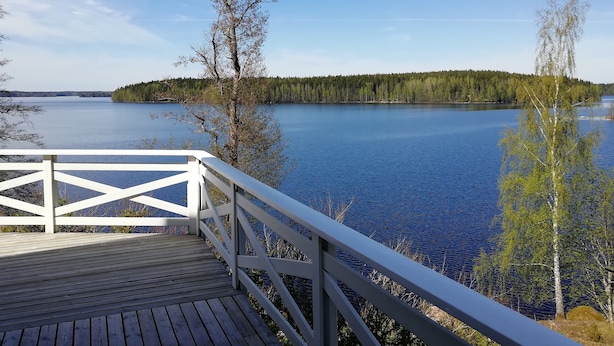 A wooden deck with a white railing overlooking a calm lake, surrounded by trees and a distant forested island. - Comfortable cottage on a beautiful lake (Uro)