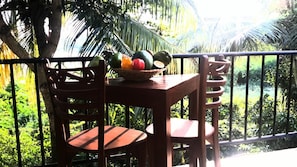 A wooden table with a fruit basket on a balcony with a metal railing, surrounded by lush greenery.