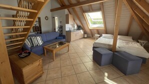 Un dormitori d'estil loft amb sostre inclinat, una escala de fusta, un sofà blau, una taula de centre de fusta i un llit amb roba de llit blanca.