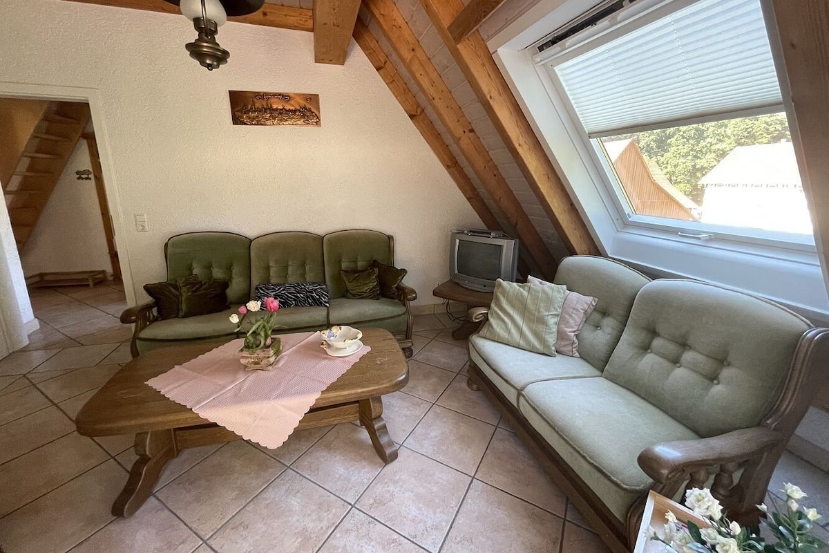 A cozy living room with a wooden ceiling, a green sofa set, a wooden coffee table with a placemat, and a television on a stand.