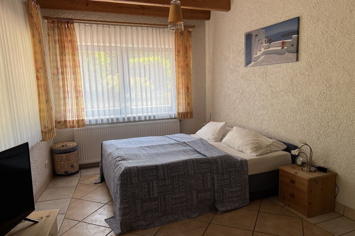 A bedroom with a bed, a bedside table, a lamp, a TV, and a window with curtains.