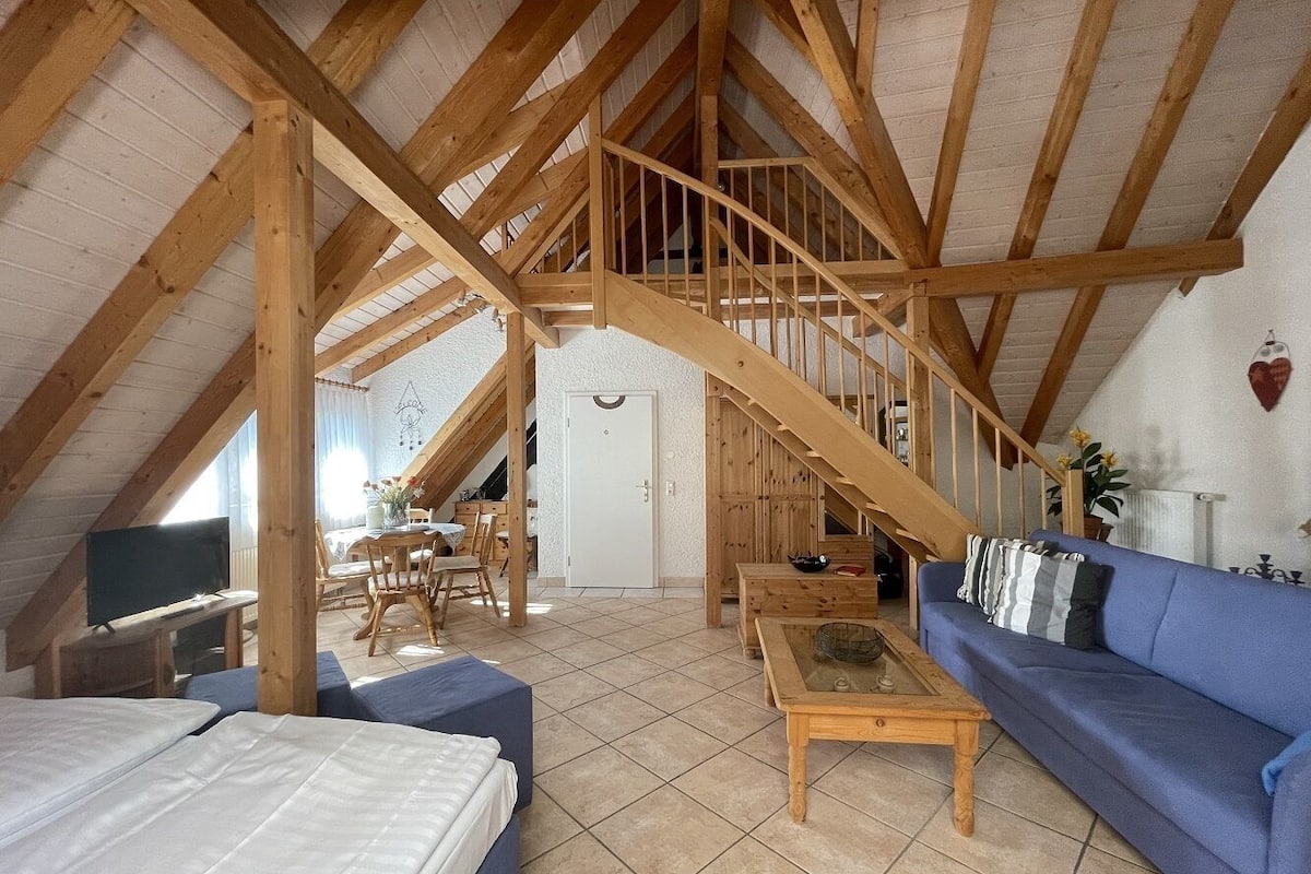 A cozy attic room with a wooden staircase, a sofa, a TV, and a dining area.