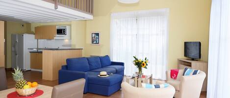 Ein Wohnzimmer mit blauem Sofa, beigefarbenen Sesseln, einem Esstisch mit Obstschale und einer sichtbaren Küche im Hintergrund.