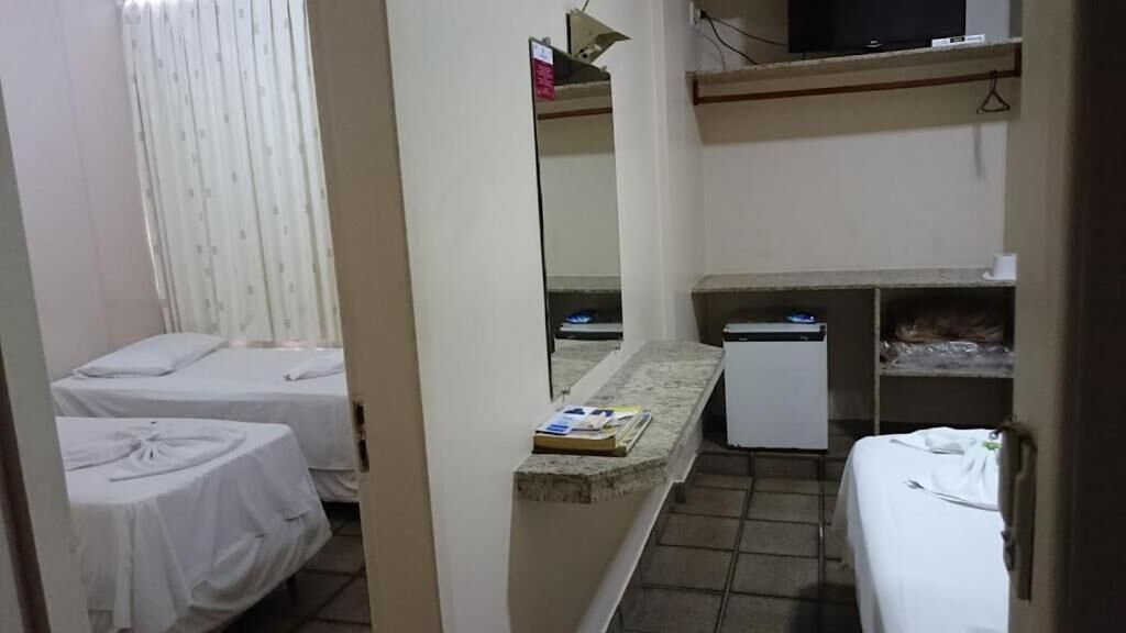 A hotel room with two beds, a small desk with a computer, a mirror, and a shelf with items.
