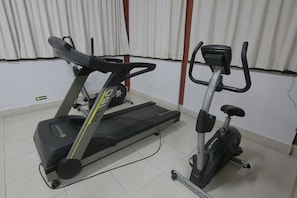 Un gimnasio en casa con dos máquinas de ejercicio, una cinta de correr y una elíptica, en una habitación con cortinas blancas y suelo de baldosas.