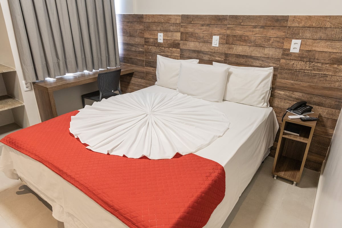 A neatly made bed with a red blanket, a wooden headboard, and a bedside table with a phone and remote control.