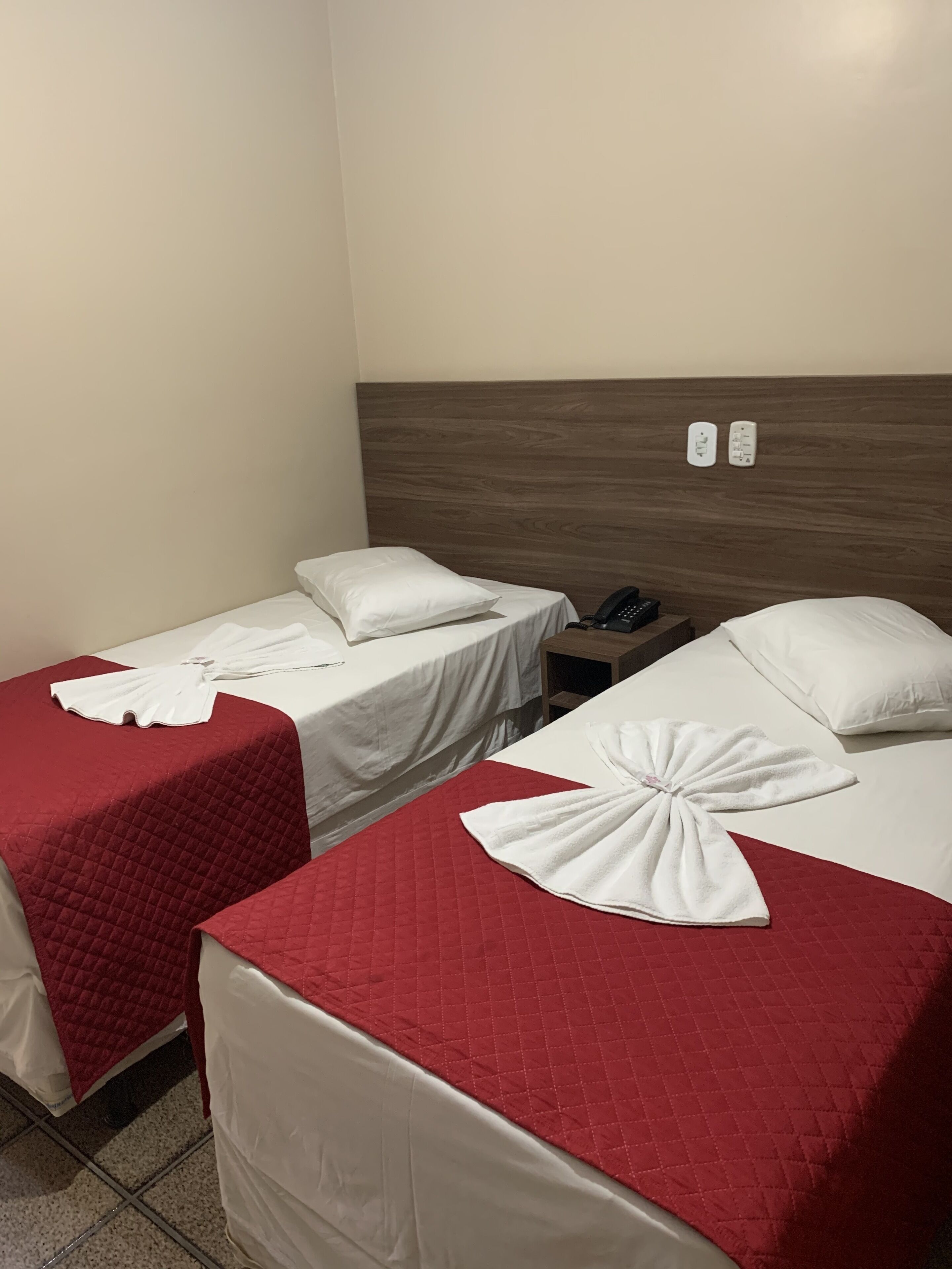 Two single beds with red quilts and white pillows, a wooden headboard, a bedside table with a phone, and two wall sockets.