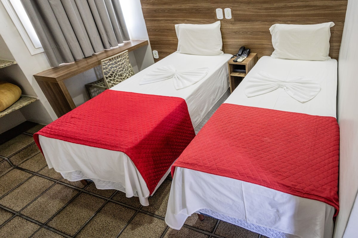 A hotel room with two beds, each with a white bedspread and a red quilted blanket. There is a wooden headboard, a nightstand with a phone, and a window with curtains.