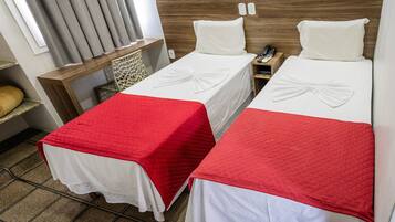 A hotel room with two beds, each with a white bedspread and a red quilted blanket. There is a wooden headboard, a nightstand with a phone, and a window with curtains.