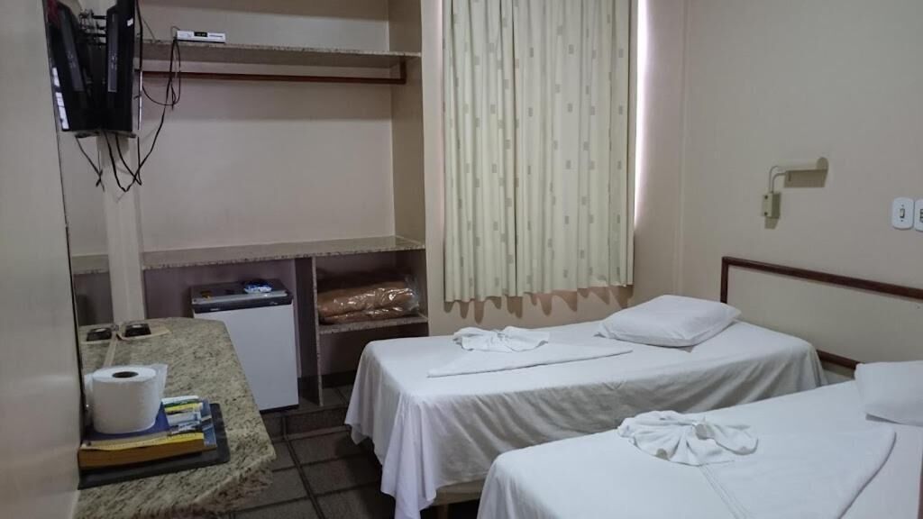 A hotel room with two single beds, a desk with a computer, a small kitchenette, and a television mounted on the wall.