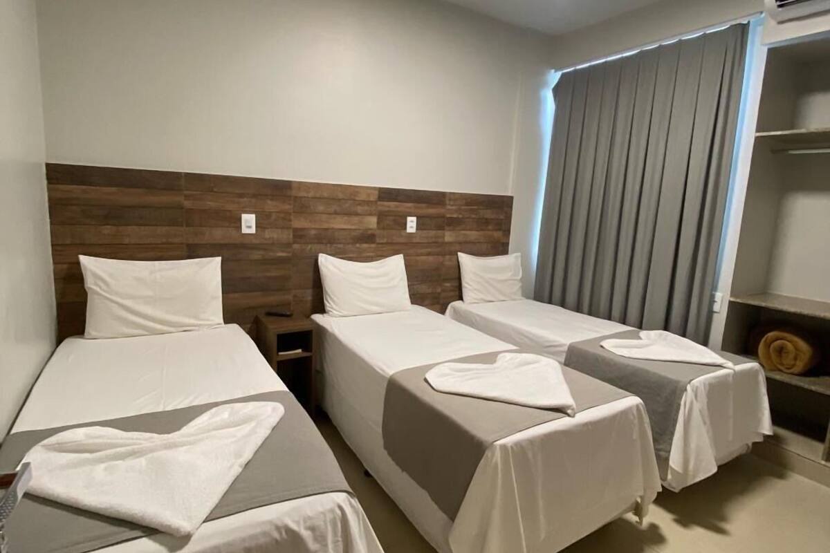 A hotel room with two single beds, a wooden headboard, a large window with curtains, and an air conditioning unit.
