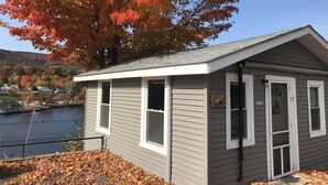 A small, single-story building with a sloped roof, white siding, and a garage door. It is located near a body of water with a dock, surrounded by trees with autumn foliage. - Cozy cottage get-away at Lake Winnipesaukee with swim access off dock (Alton)