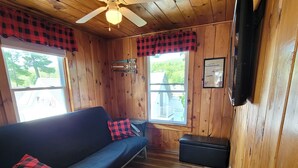 A cabin interior with a wooden floor, a sofa with a plaid pillow, a ceiling fan, and a television mounted on the wall.