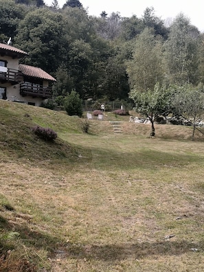 Une maison avec un toit en pente et un balcon, entourée d’une cour verdoyante avec quelques arbres et une clôture.