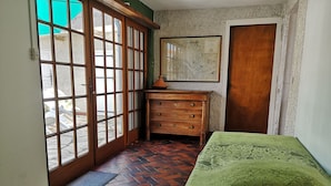 A room with a wooden dresser, a bed with a patterned bedspread, a map on the wall, and a view through a glass door.