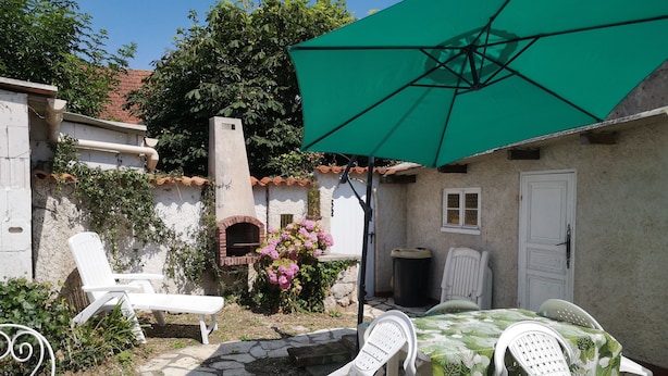 A patio with stone flooring, white plastic chairs, a table with a green tablecloth, a green umbrella, a brick oven, and a white door.