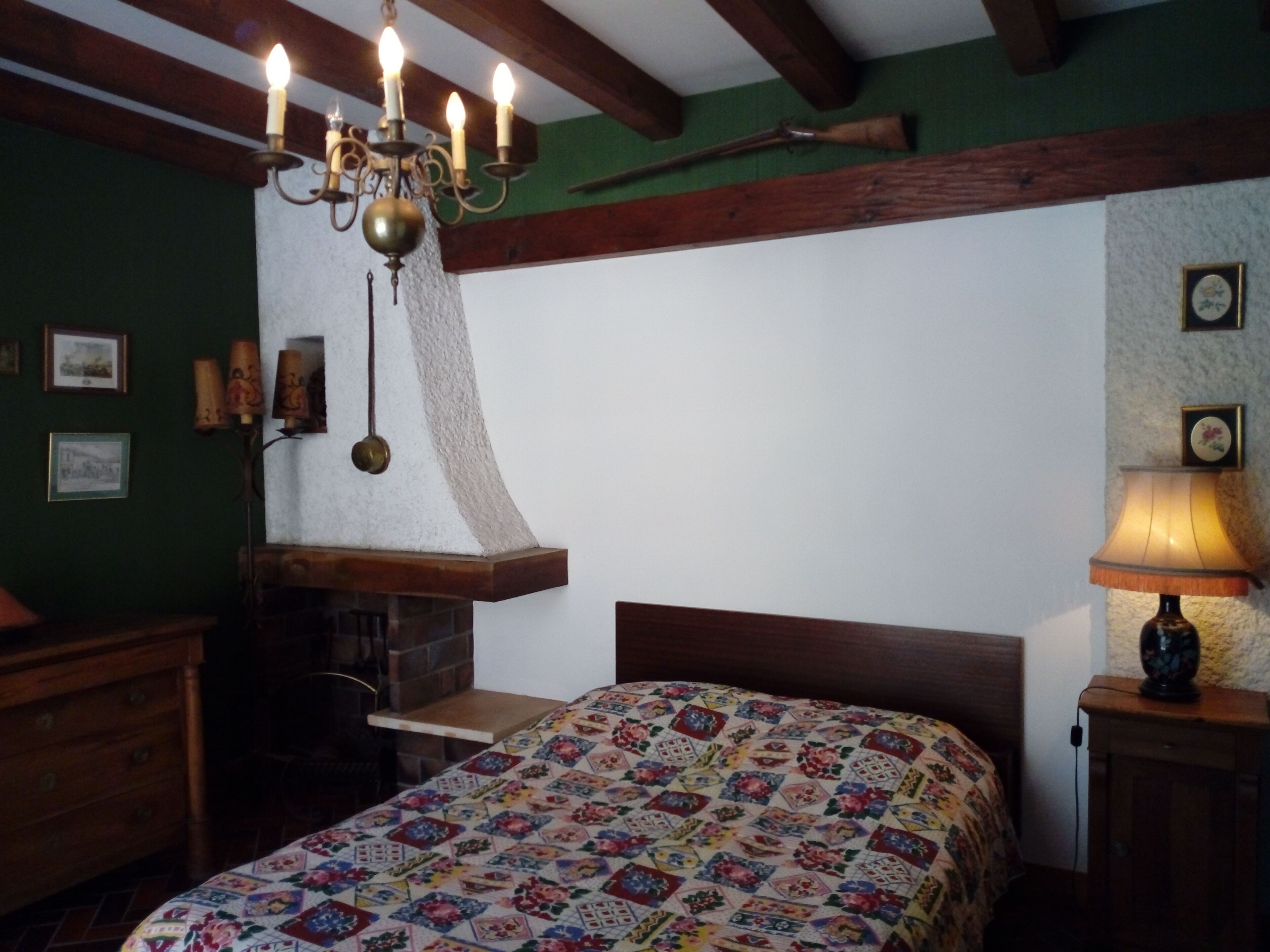 A bedroom with a wooden headboard, a quilted bedspread, a chandelier, a fireplace, and a nightstand with a lamp.