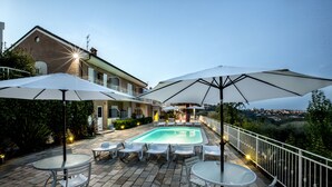 Zona de piscina con mobiliario exterior y sombrillas, rodeada por una cerca blanca y con vistas a un paisaje pintoresco.