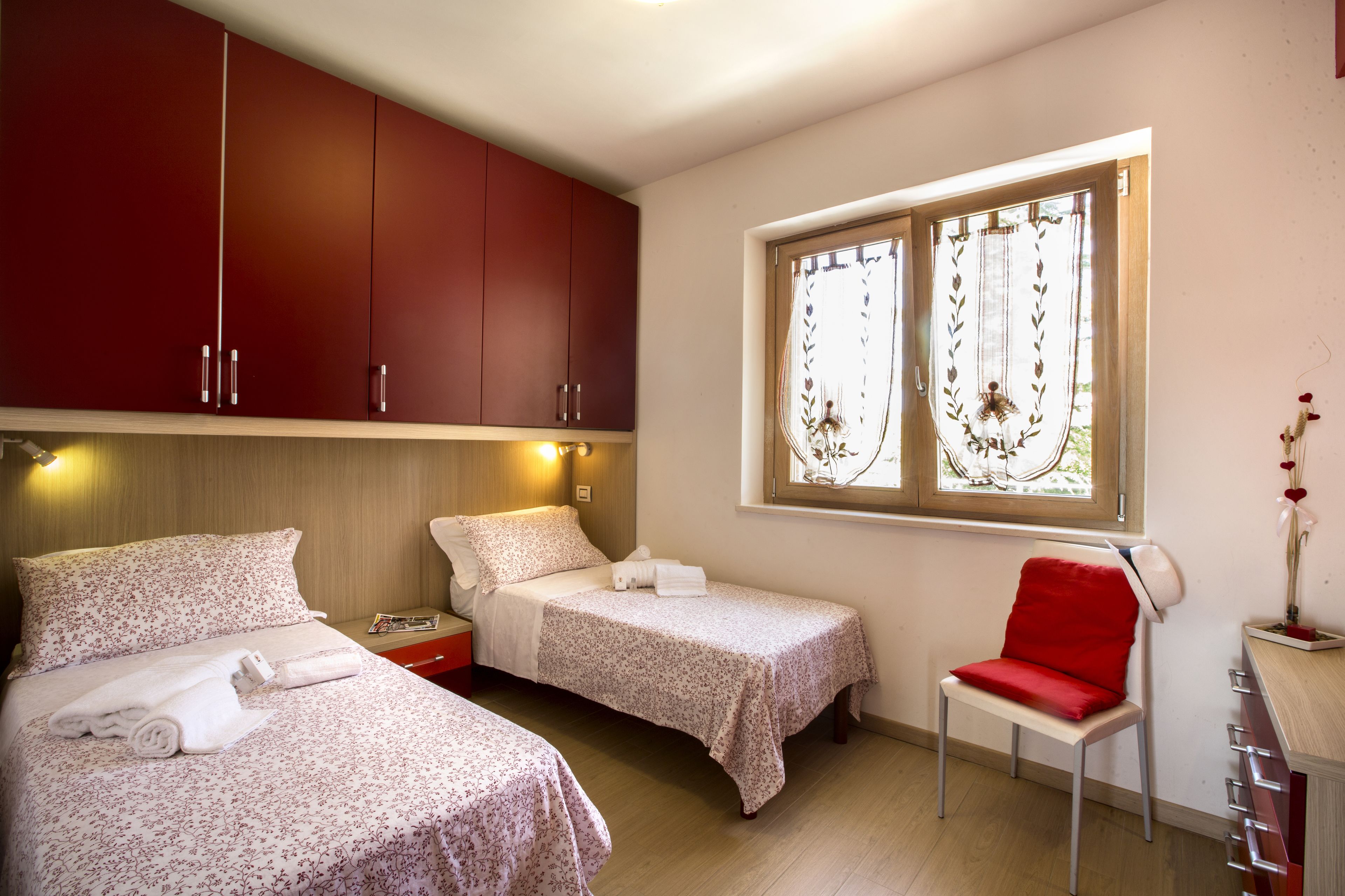 Une chambre avec deux lits, une fenêtre agrémentée de rideaux décoratifs, un fauteuil rouge et une table de chevet avec une lampe.