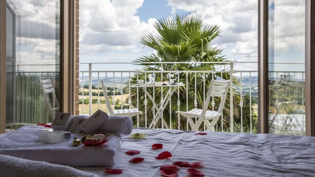 Un balcón con una mesa y sillas, una cama con colcha blanca y pétalos de rosa, y vista al campo.