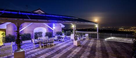 A covered outdoor patio with seating and planters, a brick floor, and a view of the city at night.