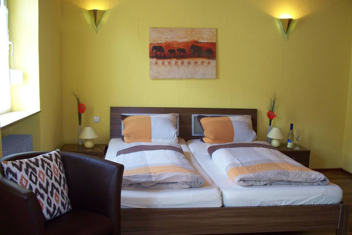 A double bed with a wooden headboard, two bedside tables with lamps, a chair with a patterned cushion, and a wall painting of elephants.