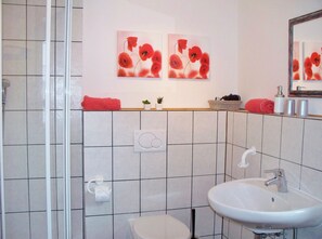 Baño con lavamanos blanco, inodoro y zona de ducha. Hay dos toallas rojas sobre el mueble y un espejo sobre el lavamanos.
