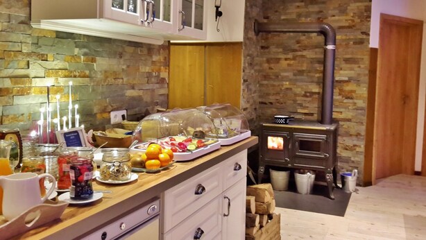 A kitchen with a stone wall, a wood-burning stove, and a breakfast bar with fruit and condiments. - Pension  Zum Stern (Freudenberg)