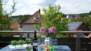 A table with wine bottles, glasses, and heart-shaped containers on a balcony overlooking a residential area with solar panels and houses. - Pension  Zum Stern (Freudenberg)