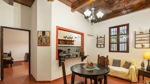 Een woonkamer met een eettafel, stoelen, een bank, een kroonluchter en een keukenhoek met planken en een raam.