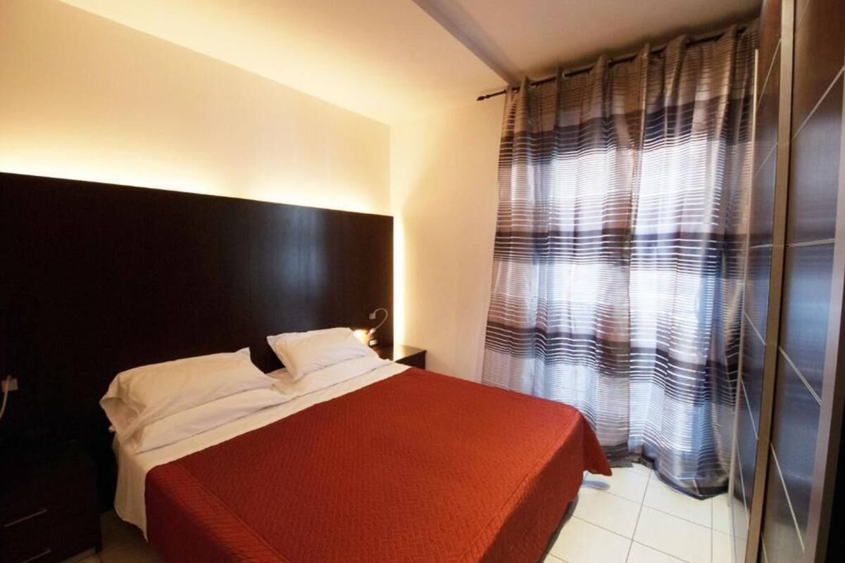 Una cama individual con una colcha roja en una habitación con cabecera oscura y cortina a rayas.