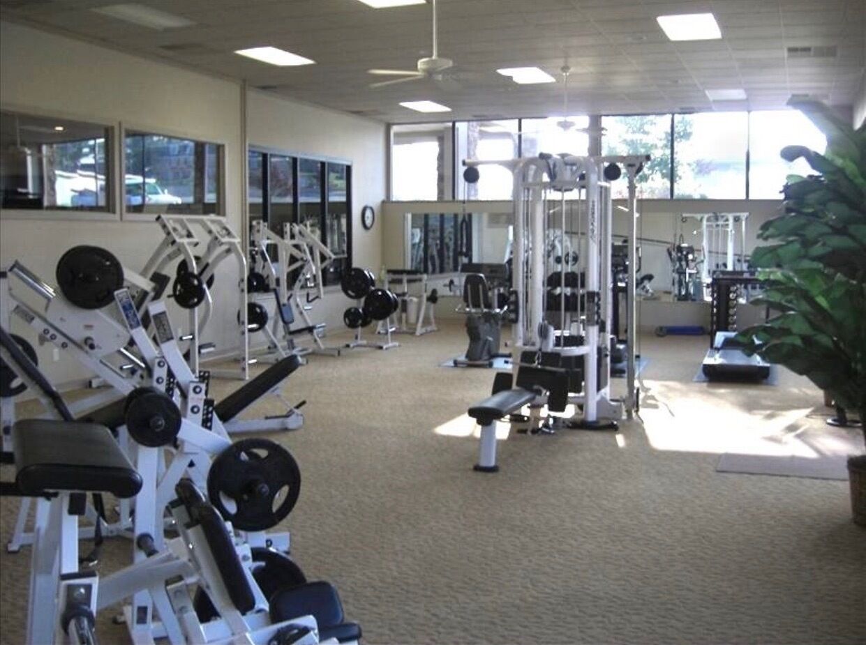 Une salle de sport équipée de divers appareils d’exercice, dotée de grandes fenêtres et d’un ventilateur de plafond.