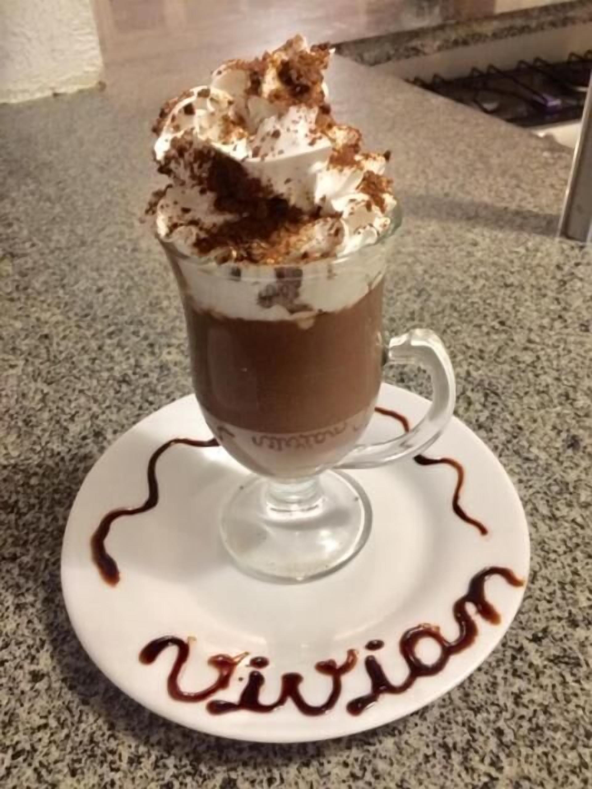 Una tazza di cioccolata calda con panna montata e scaglie di cioccolato su un piattino con la scritta 'Vivian' in cioccolato.