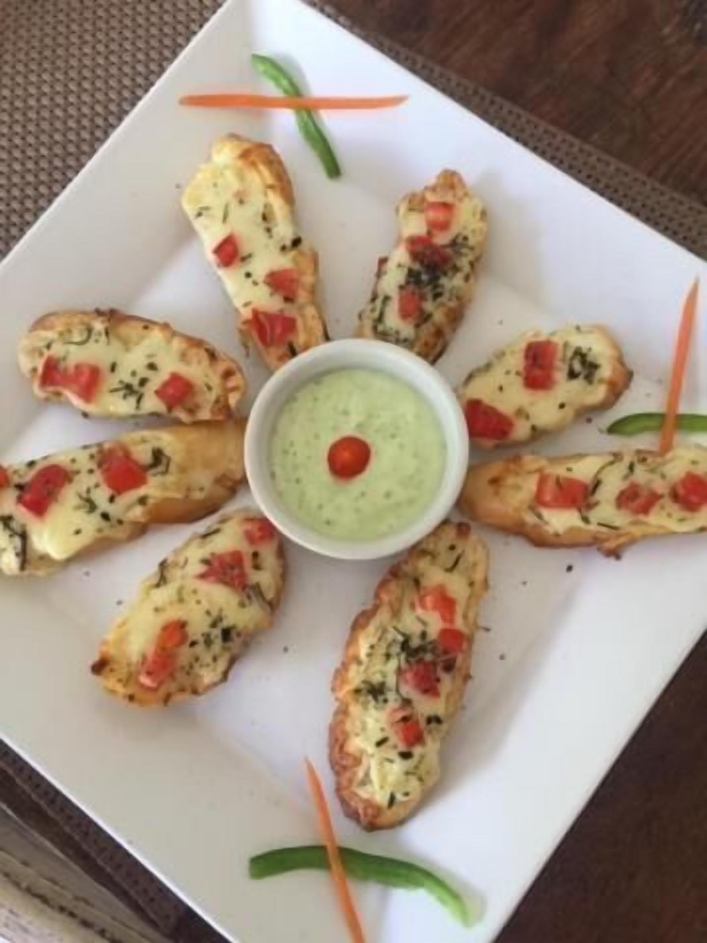 A white plate with a central bowl of green dip surrounded by garlic breadsticks with red peppers.