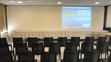 Et mødelokale med projektorskærm der viser et softwareinterface, et langt bord og rækker af stole.