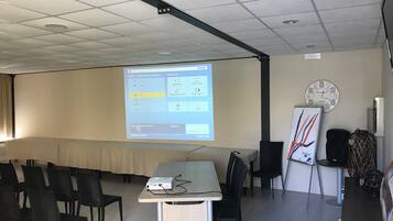 Uma sala de conferências com tela de projetor exibindo uma apresentação, um quadro branco e um relógio na parede.